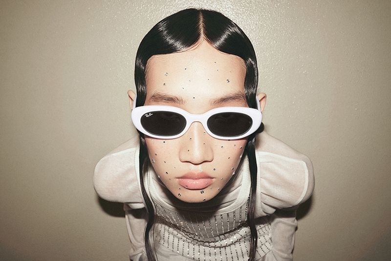 Close-up portrait of a person wearing white sunglasses and a sheer top, against a neutral wall, with applied silver glitter.