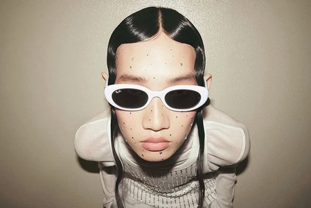 Close-up portrait of a model wearing white oval sunglasses and rhinestone facial gems against a neutral background.