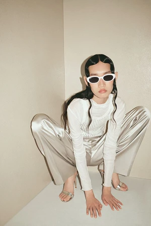 Woman wearing white oval sunglasses and satin pants crouching in a neutral corner for Ray-Ban Holiday Campaign 2024.