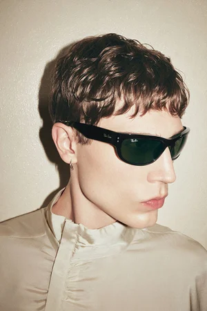 Close-up profile portrait of a man wearing black sunglasses and a cream high-collar shirt against a textured wall.