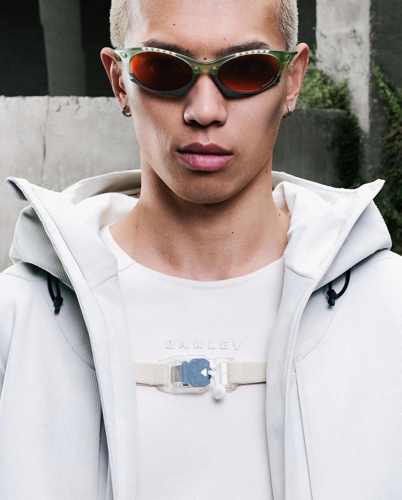 Close-up of an adult male with short blonde hair, wearing green sunglasses and a white jacket, standing outdoors against a stone wall.