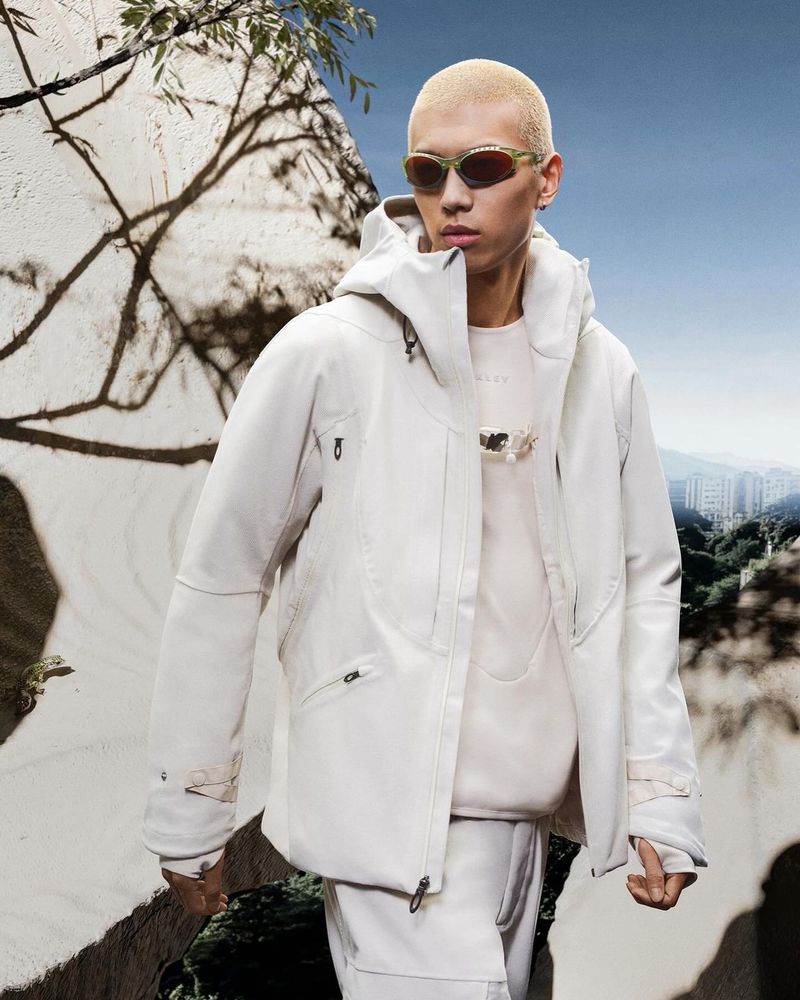 An adult male model wearing sunglasses and a light-colored hooded jacket stands outside against a background of trees and a cityscape.