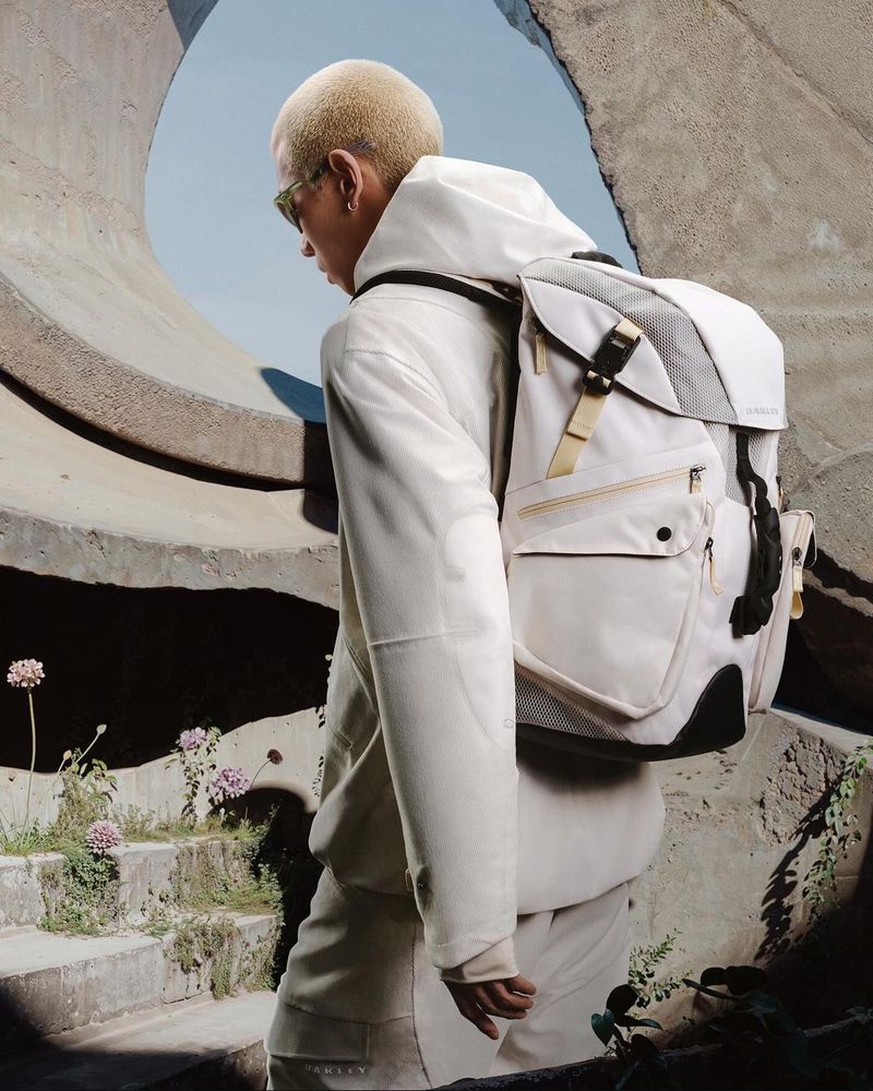Person with short blonde hair and sunglasses wearing a backpack in front of a concrete architectural structure with plants and stairs.
