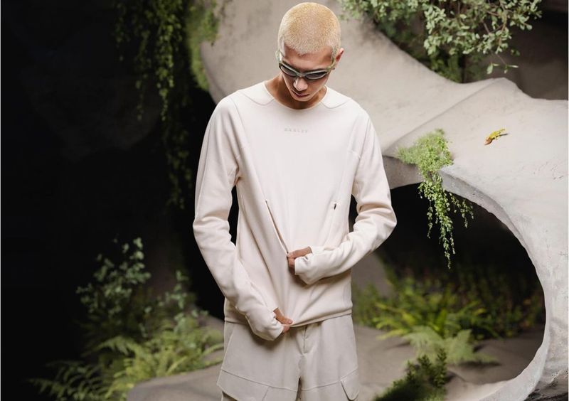 A light-skinned male model with a shaved head wears light-colored clothing in a neutral, outdoor setting with lush greenery and rock formations.