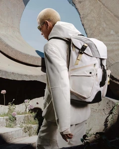 Model wearing a white hoodie and carrying a white backpack outdoors near concrete architectural structures