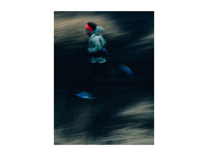 Motion blur image of an adult male jogging on a dark surface wearing a light jacket and red headwrap. Shot with a slow shutter speed.