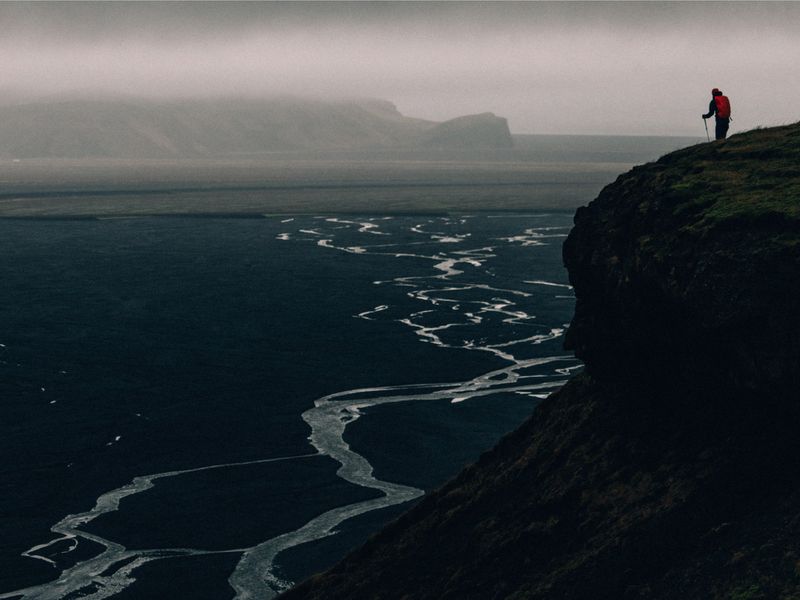 A lone hiker stands on a cliff overlooking a dark body of water with winding, light-colored patterns, set against a foggy landscape.