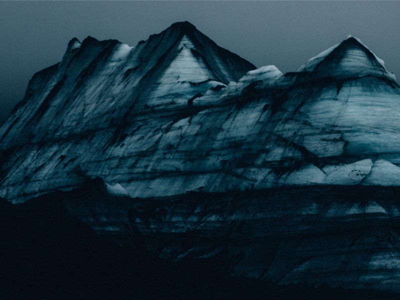 A striking landscape features a rugged, snow-covered mountain range under an overcast sky, evoking a sense of stark beauty and isolation.