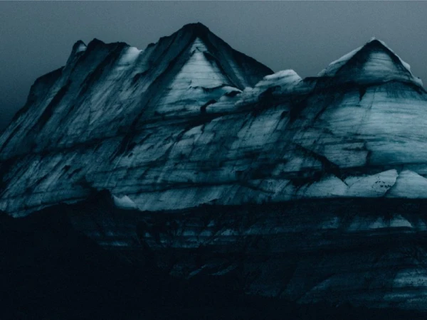 A striking landscape features a rugged, snow-covered mountain range under an overcast sky, evoking a sense of stark beauty and isolation.