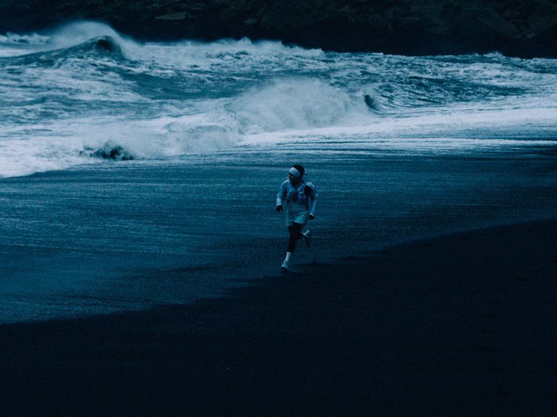 Runner in athletic wear runs on a black sand beach alongside white waves in the ocean. The image exhibits a monochromatic blue color grading.