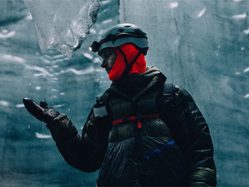 Man in helmet and down jacket explores icy cavern, holds out gloved hand to catch dripping water in frigid, gray environment.