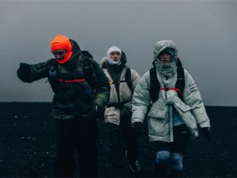 Three hikers in heavy insulated outerwear and balaclavas walk across dark terrain under an overcast, foggy sky.