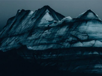 Dark, dramatic mountain peaks with layered ice, snow, and volcanic sediment under an overcast deep blue sky.
