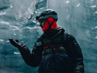 Ice climber wearing a helmet, bright orange balaclava, and puffer jacket catching water dripping from melting glacier ice.