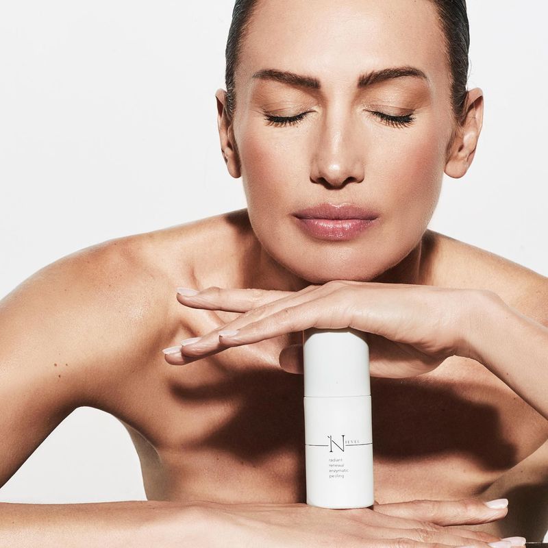 Adult woman with closed eyes resting her face on her hands, next to a white cosmetic bottle with text, on a white background.