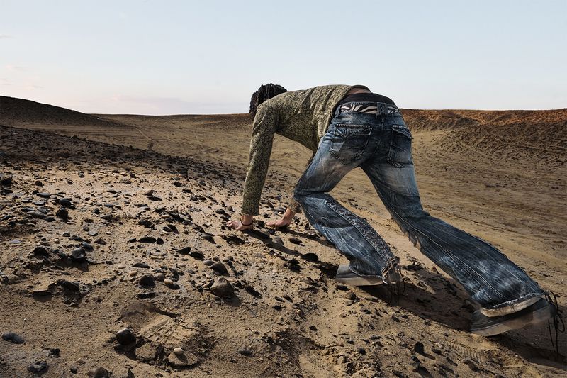 A person crawls on hands and knees up a sandy hill in an arid landscape under a bright sky, viewed from behind.