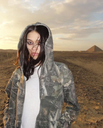 Young woman in a camouflage hoodie posing outdoors with a pyramid in the background during a sunset, creating an editorial style image.