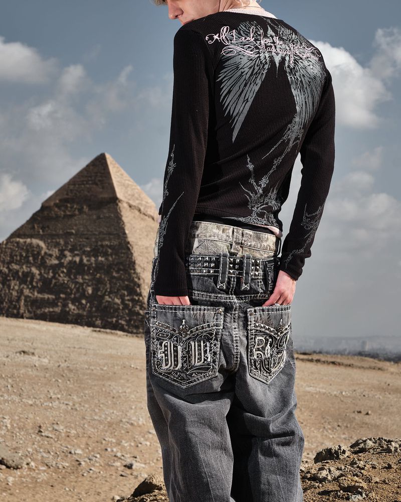 A person wearing a long-sleeve shirt and jeans poses in a desert environment with a large pyramid in the background.