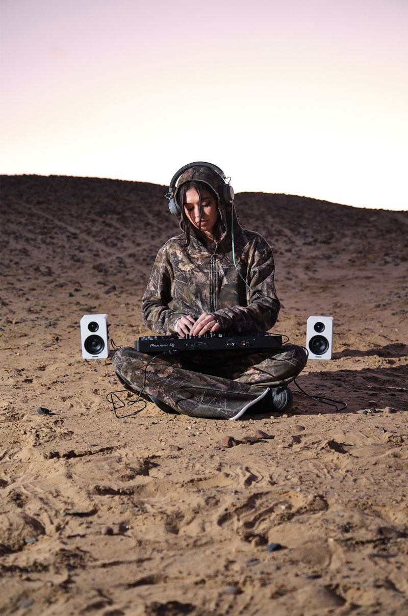 Woman in hooded camo jacket sits cross-legged in sand playing music on a MIDI controller with two speakers, wearing headphones.