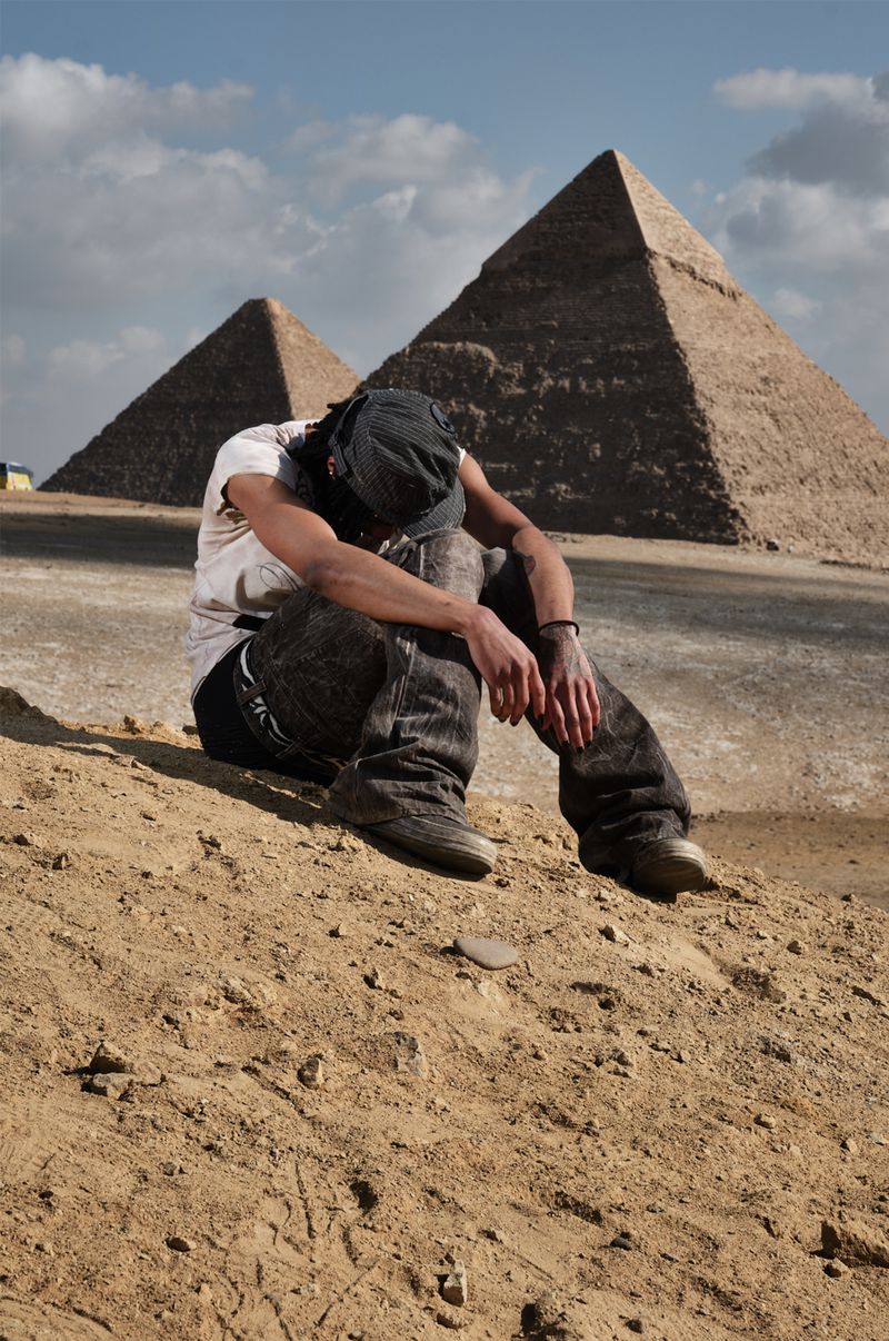 Man sits on sand in front of pyramids in Egypt, wearing cap, t-shirt, jeans and shoes, with head down and hands on knees, under blue sky.