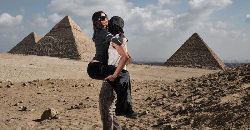 Adult male lifts woman outdoors near pyramids in desert landscape under cloudy sky in medium shot, embracing travel and exploration.
