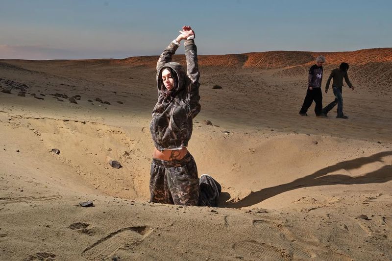 A woman in a camouflage hoodie and pants kneels in a sandy pit in the desert with her arms raised, illuminated by warm sunlight.