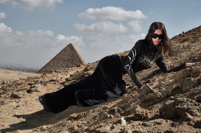 A woman in a black top and long skirt poses on a rocky hill in front of a pyramid on a clear day, wearing sunglasses.