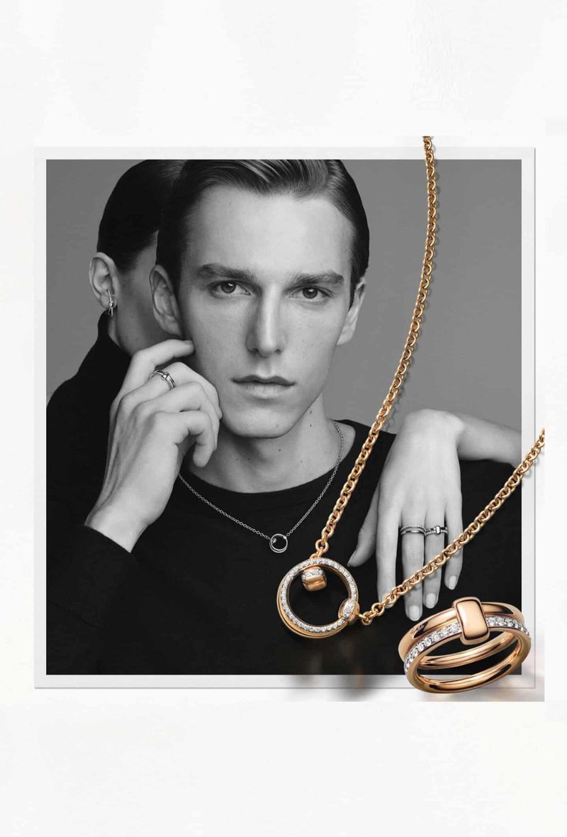 Portrait of a young adult male model in black top, wearing a necklace with gold chain and ring-shaped pendant, gold rings are prominent.