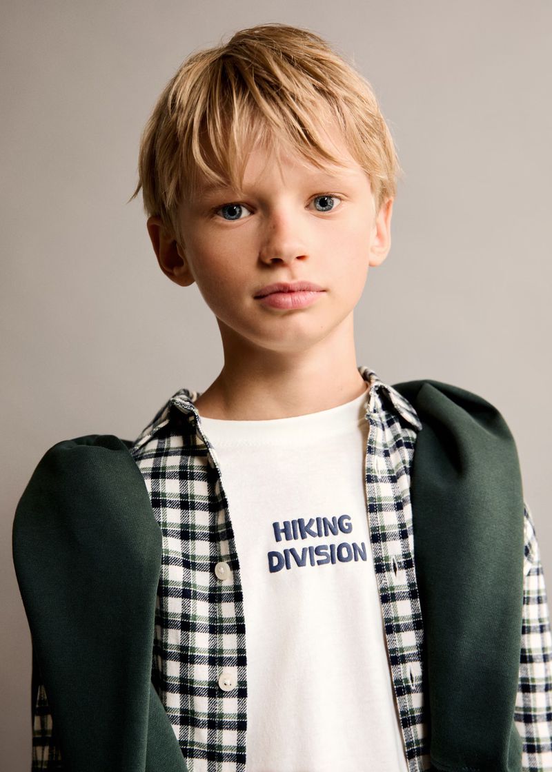 Young boy in white t-shirt and plaid shirt, posing indoors. He is also wearing a green sweater over his shoulders.