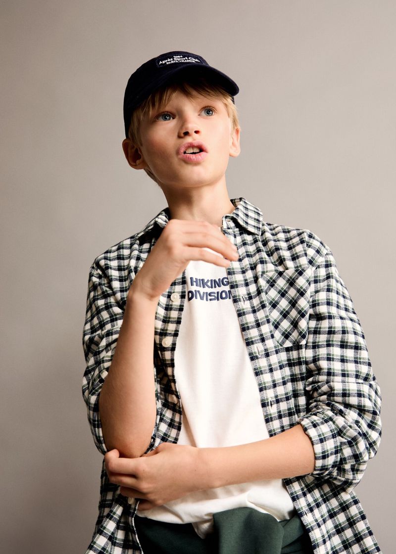 A young boy in casual fashion, wearing a plaid shirt, t-shirt, and baseball cap, poses in a medium shot against a neutral background.