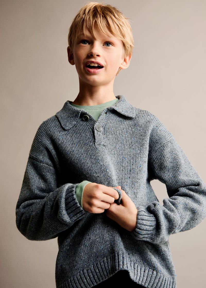 Blond child in a blue, buttoned, collared sweater and green t-shirt poses in a medium indoor shot against a plain backdrop.