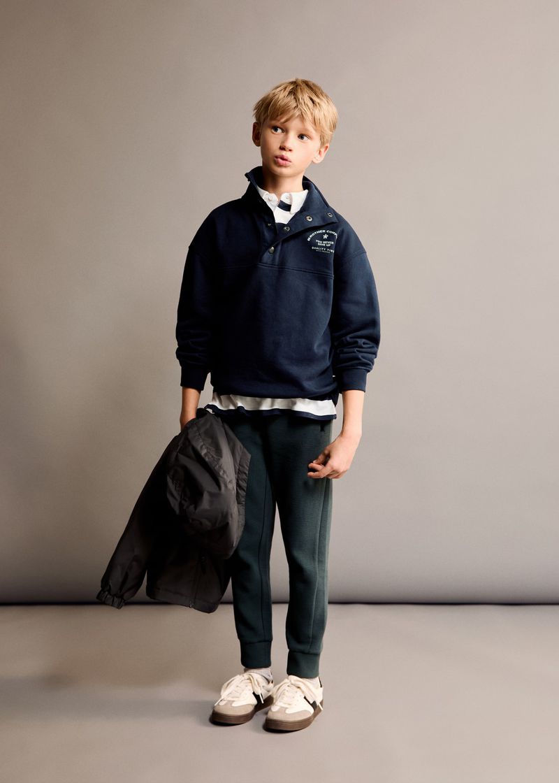 Boy in navy buttoned sweatshirt and green sweatpants stands in a photo studio with a jacket slung over his arm. Neutral background.