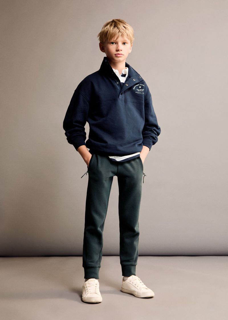 Young blond boy poses indoors wearing navy buttoned sweatshirt, dark green jogger pants, and off-white sneakers against a gray background.