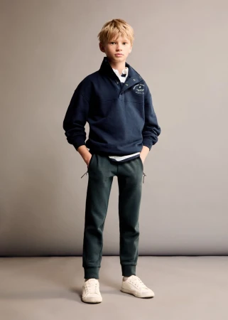Young blond boy poses indoors wearing navy buttoned sweatshirt, dark green jogger pants, and off-white sneakers against a gray background.