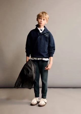 A young boy standing against a plain grey backdrop wearing a navy blue sweatshirt and dark sweatpants, holding a dark jacket.