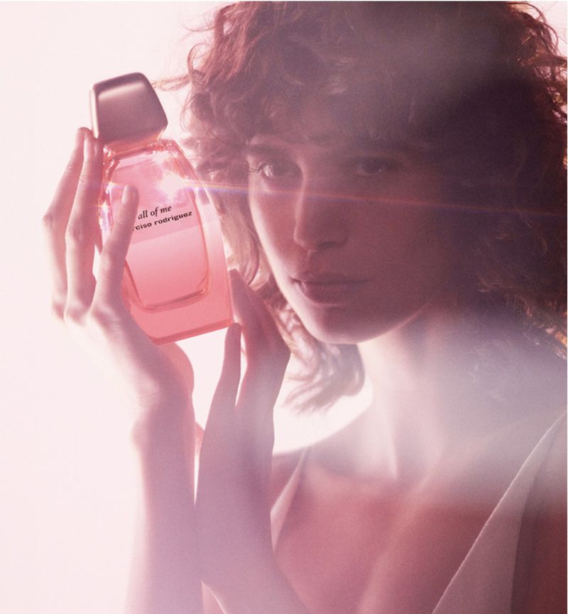A woman holds a pink bottle of 'all of me' Narciso Rodriguez perfume in a soft-focus studio shot, with gentle lighting and a modern aesthetic.