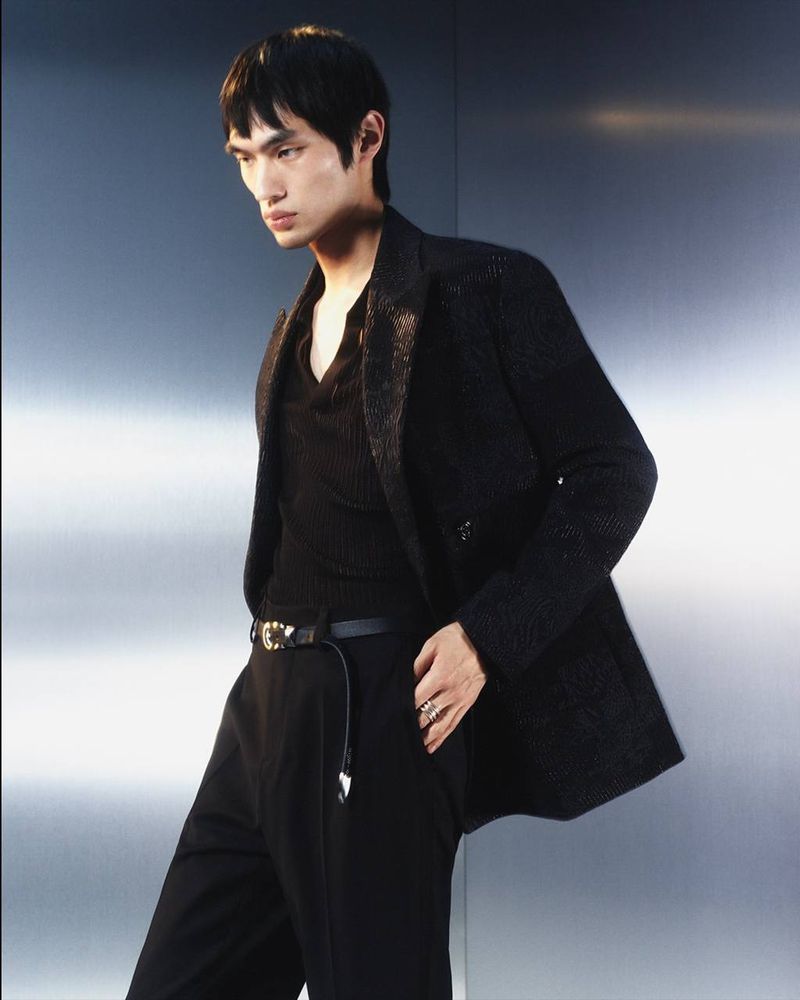 Asian male model poses in black suit jacket and pants against a gray background in a fashion portrait with soft studio lighting.