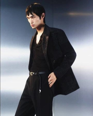 Asian male model poses in black suit jacket and pants against a gray background in a fashion portrait with soft studio lighting.