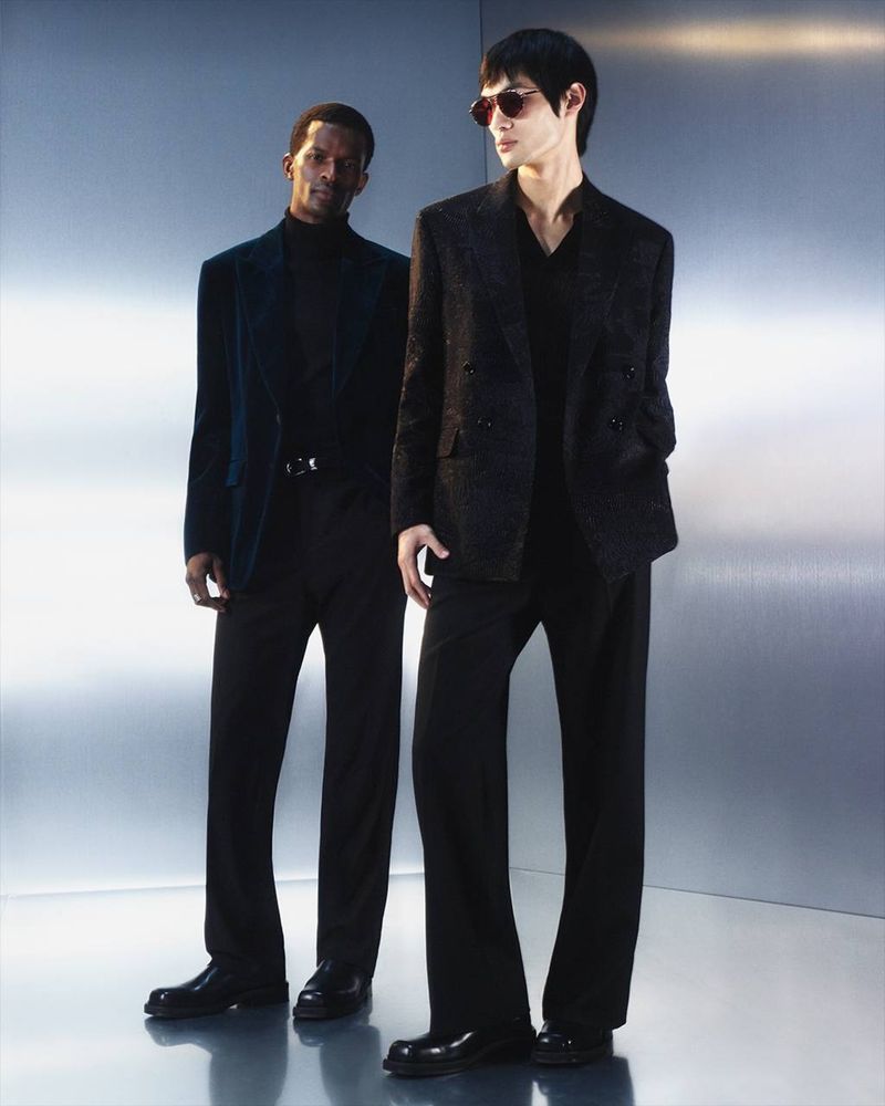 Two male fashion models in a photo studio. One wears a blue blazer with a black turtleneck; the other has a black suit and sunglasses.