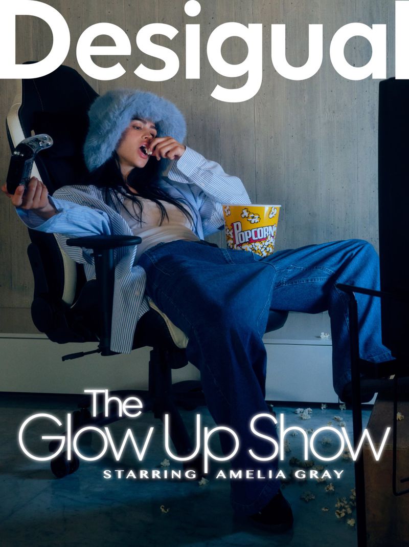 Woman in blue fuzzy hat and jeans sits in gaming chair eating popcorn and holding game controller in an indoor setting with TV.