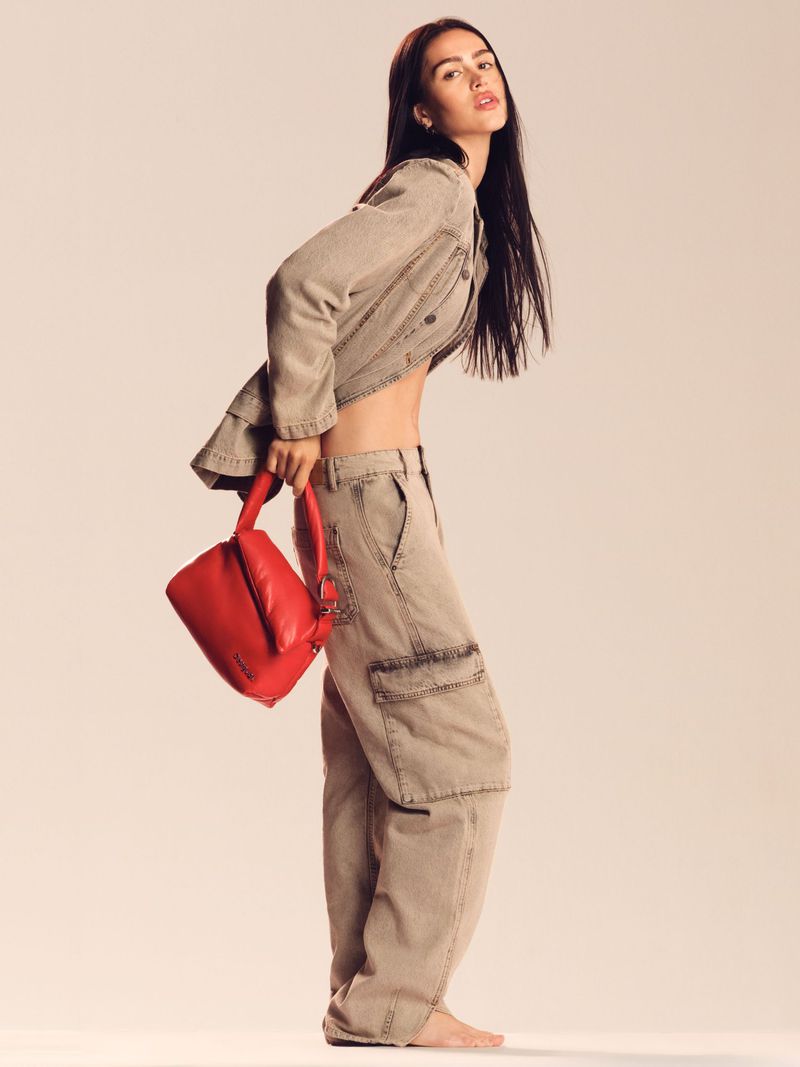A woman poses in a tan denim jacket and pants, holding a red purse, against a light background. The pose is three-quarters, showcasing the outfit.