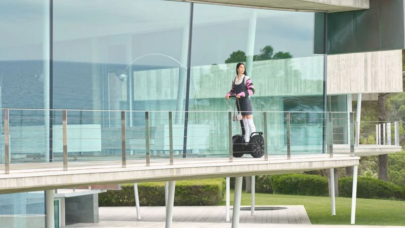 A woman riding a black Segway on a modern building's walkway with glass walls, showcasing architecture, transportation, and outdoor lifestyle.
