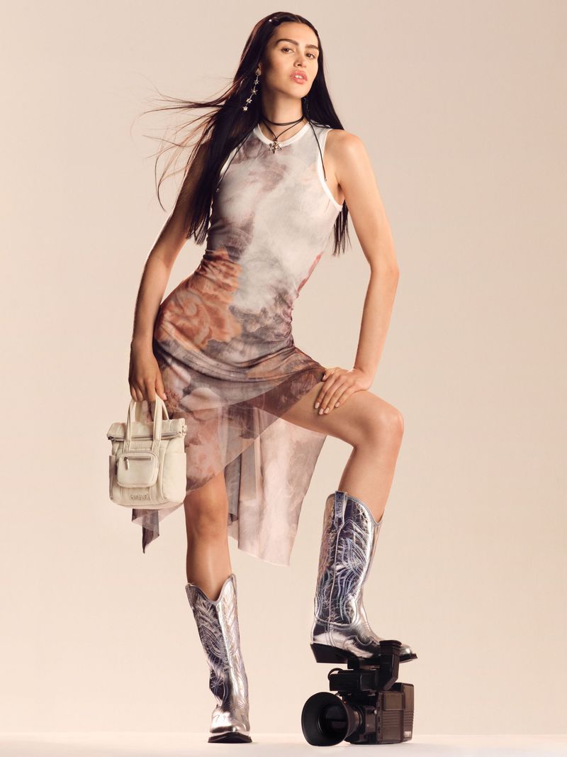 A woman in a patterned dress and cowboy boots poses with a vintage camera and purse, against a neutral backdrop in a fashion editorial.