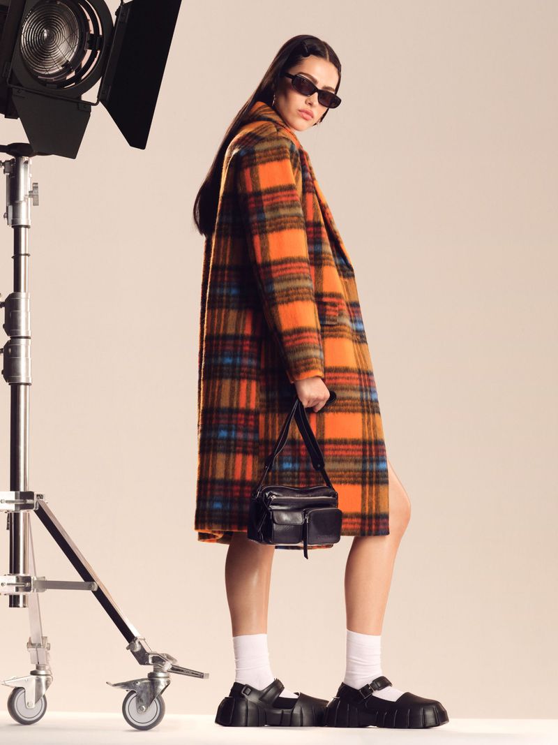 Woman in orange plaid coat holding black purse, wearing black sunglasses and chunky shoes stands in front of a photo studio light.