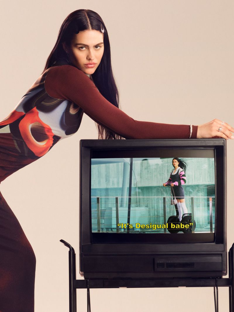 Adult female posing next to a vintage television in a studio setting, wearing a brown top and patterned dress with long dark hair.