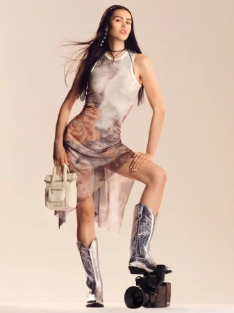 Amelia Gray wearing a printed sheer sleeveless dress and silver cowboy boots resting one foot on a camcorder against a studio backdrop.