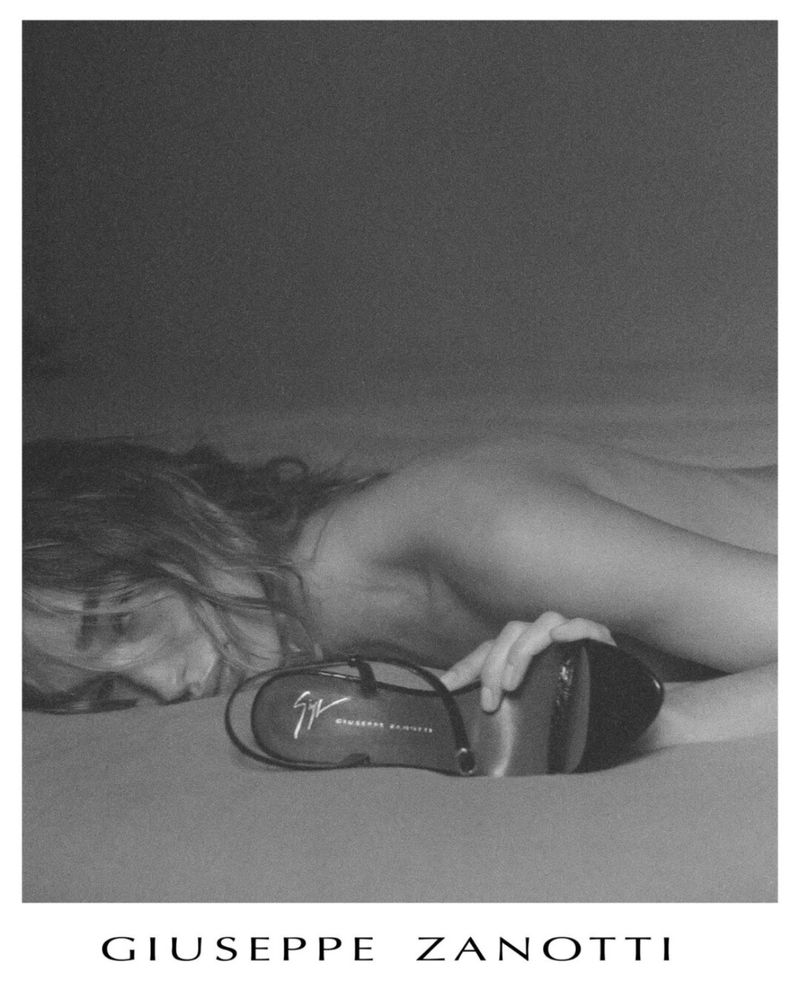Black-and-white studio product shot featuring a woman laying on a bed next to a Giuseppe Zanotti high-heeled sandal.