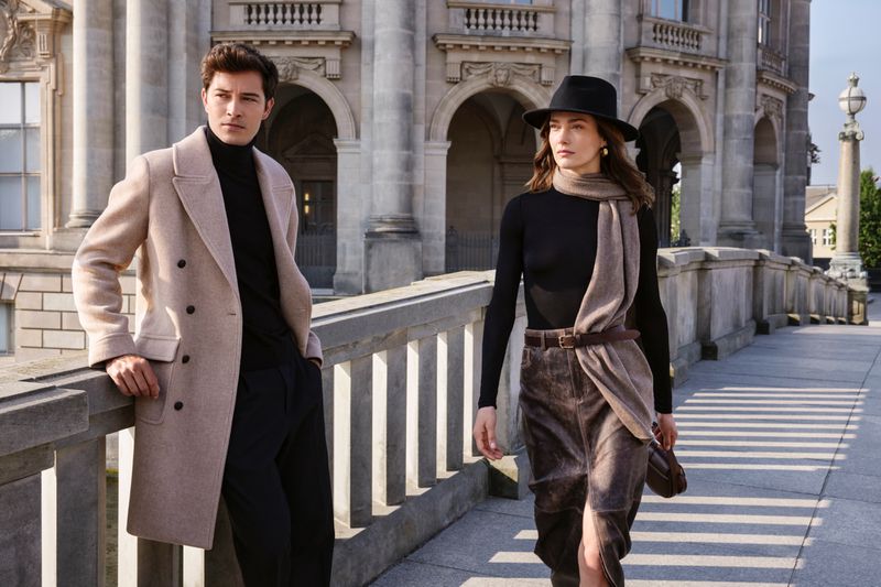 Two fashion models pose on a bridge in neutral-toned clothing with a building backdrop, showcasing elegant outerwear and modern style.