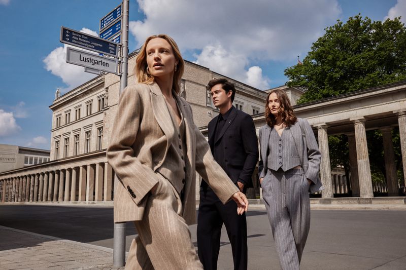 Three fashion models in stylish suits pose in an outdoor urban setting, with historic architecture and blue sky visible in the background.
