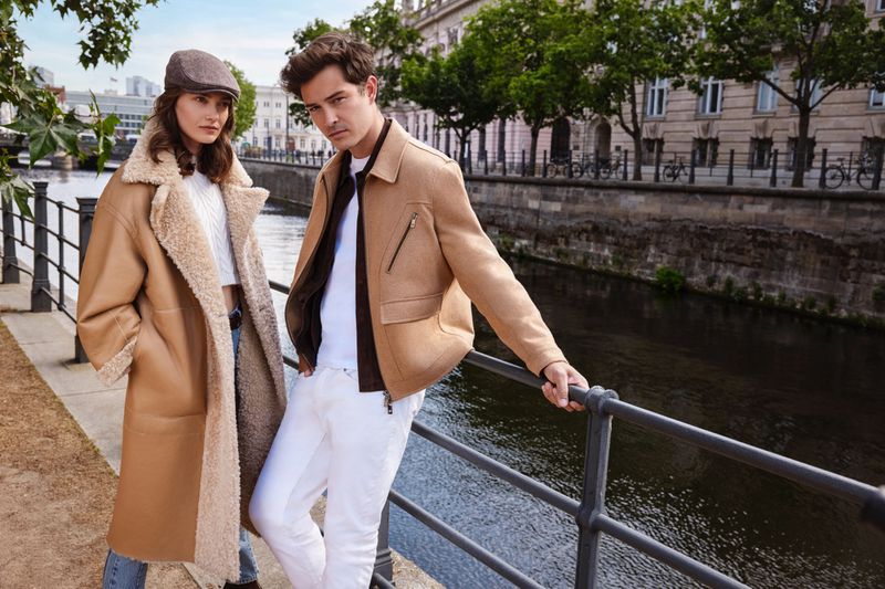 A stylish couple poses outdoors on a city street in Europe; the woman wears a long coat, and the man a short jacket, both in tan.